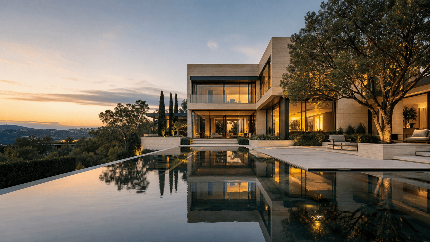 Mansão contemporânea de dois pavimentos com fachada de pedra clara e amplos vidros iluminados ao entardecer, refletida em uma piscina de borda infinita, cercada por árvores e vegetação verde.